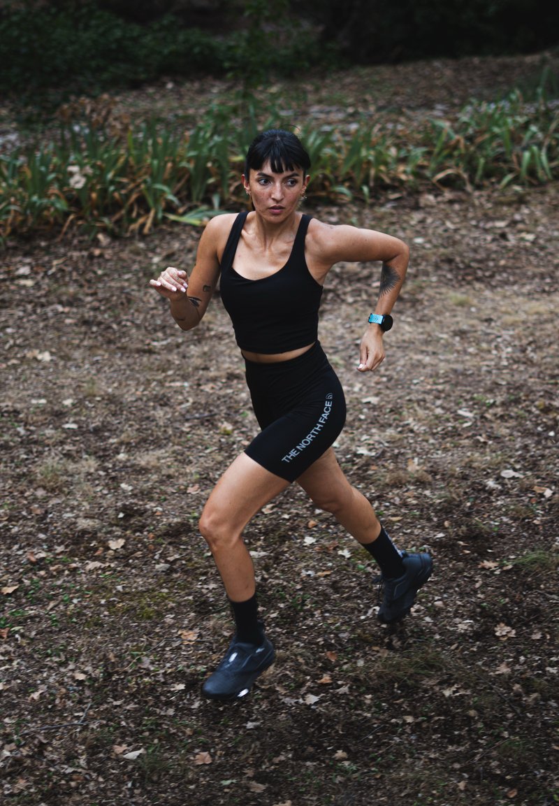 Woman wearing black sportswear and a fitness watch running on a dirt path in a wooded outdoor area.