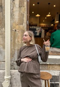 Brun texturerad överdel och matchande byxor, hålls av en person med ett glas kall dryck. Avslappnad miljö med en stenvägg och trädetaljer.