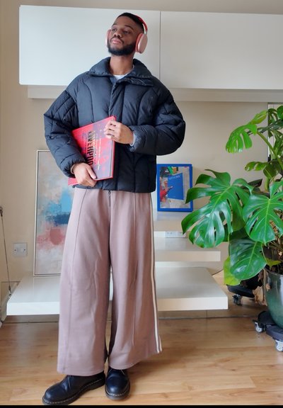 Hombre con chaqueta acolchada negra, pantalones anchos beige y auriculares rosas sosteniendo un disco rojo en una sala de estar moderna.