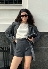 Abrigo de lana gris sobre un suéter blanco, combinado con pantalones cortos grises de cintura alta. Accesorios incluyen gafas de sol negras y un pequeño bolso negro.