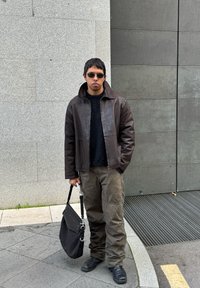 Καφέ δερμάτινο μπουφάν πάνω από ένα μαύρο πλεκτό πουλόβερ, ελαιοπράσινα παντελόνια και μαύρες μπότες. Κρατάει μια μαύρη τσάντα με ασημί λουρί.