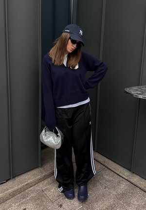 Joven mujer con suéter azul marino, pantalones deportivos negros, gorra azul marino y gafas de sol, sosteniendo un bolso plateado, de pie frente a paneles de pared oscuros.