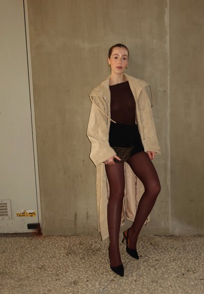 Trench coat de ante beige, top marrón, pantalones cortos negros, medias marrones y tacones negros. Sosteniendo un bolso de mano de estampado marrón. Fondo sencillo.