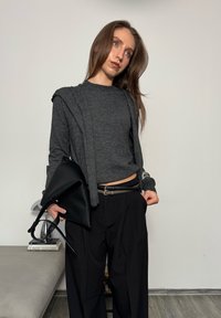 Femme vêtue d'un pull gris foncé et d'un pantalon noir, tenant un sac à main en cuir noir, debout à l'intérieur avec des livres et une lampe en arrière-plan.