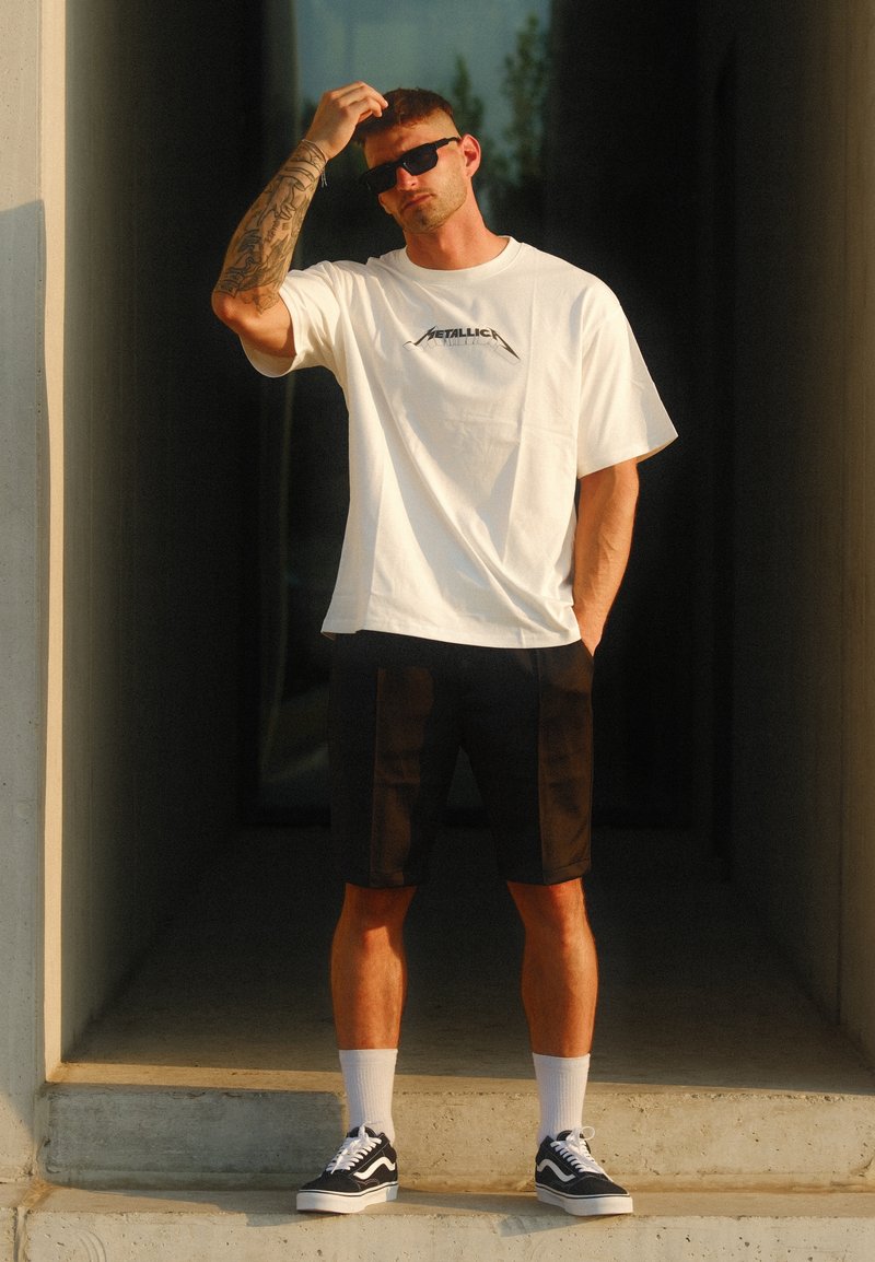 Λευκό κοντομάνικο πουκάμισο με μαύρο λογότυπο, μαύρες βερμούδες και μαύρα και λευκά αθλητικά παπούτσια. Μοντέλο που φοράει γυαλιά ηλίου σε εξωτερικό χώρο.