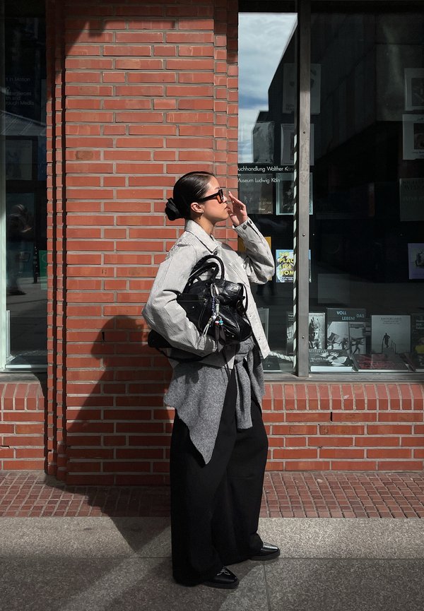 Vrouw met zonnebril, die een zwarte tas vasthoudt, staat tegen een bakstenen muur vlak bij een glazen raam met boeken.
