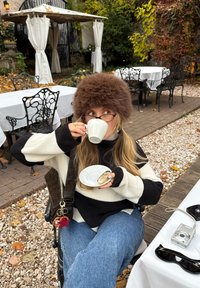 Kvinde i pels hat og stribet sweater sidder på en udendørs café og drikker te, med et kamera og solbriller på bordet ved siden af.