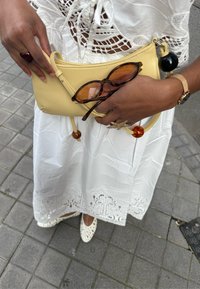 Borsa in pelle gialla con dettagli, occhiali da sole marroni e un bracciale dorato, indossati con un vestito bianco ricamato e scarpe decorative.