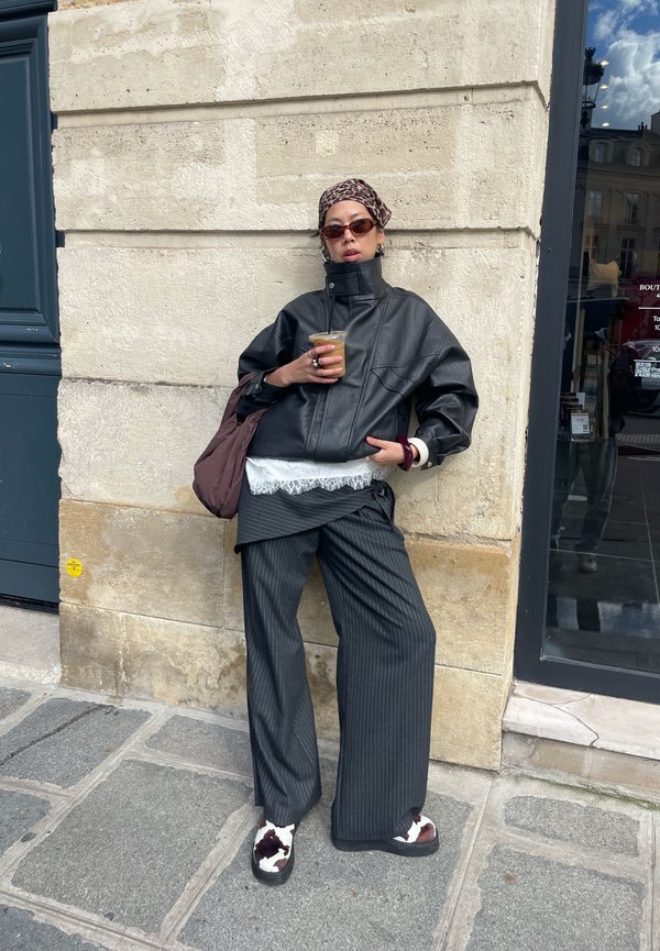 Személy kőfalnak dőlve, fekete bőrdzsekit, csíkos, széles szárú nadrágot, mintás fejkendőt és sötét napszemüveget visel, kezében jeges kávét tart.