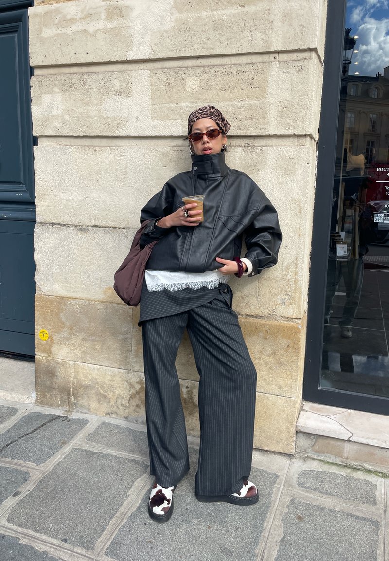 Persoană sprijinită de un zid de piatră, purtând o jachetă neagră de piele, pantaloni largi cu dungi, eșarfă cu model, ochelari de soare închiși la culoare, ținând un pahar cu cafea rece.