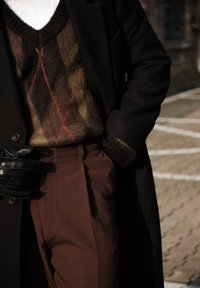 Personne portant un manteau noir, un pull argyle marron, un pantalon marron, et des gants en cuir noir, debout à l'extérieur sur un sol pavé.