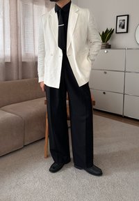 Homme portant un blazer blanc, une chemise noire, une cravate noire et un pantalon noir à jambes larges, debout dans un salon moderne avec un canapé beige et une armoire blanche.