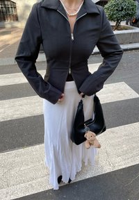 Giacca nera aderente con zip e colletto, abbinata a una gonna bianca fluente. Presenta una borsa nera con un piccolo charm a forma di orsetto.