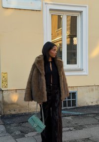 Manteau en fausse fourrure marron avec un large col, haut noir découpé, pantalon noir brillant taille haute et sac matelassé vert menthe.