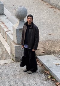 Ένα άτομο με καφέ δερμάτινο μπουφάν στέκεται σε ένα λιθόστρωτο μονοπάτι, κρατώντας μια μαύρη τσάντα, φορώντας χαλαρούς μαύρους παντελόνες και μια υφασμάτινη μπλούζα από κάτω.