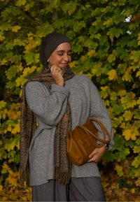 Pull gris oversize, écharpe marron à motifs avec franges, sac à main marron, pantalon gris, sur un fond de feuilles vertes et jaunes.