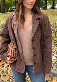 Brązowa zamszowa kurtka z zapięciem na guziki, nałożona na jasnobrązowy sweter w serek. Trzyma teksturowaną brązową kopertówkę, z dżinsami widocznymi w tle.