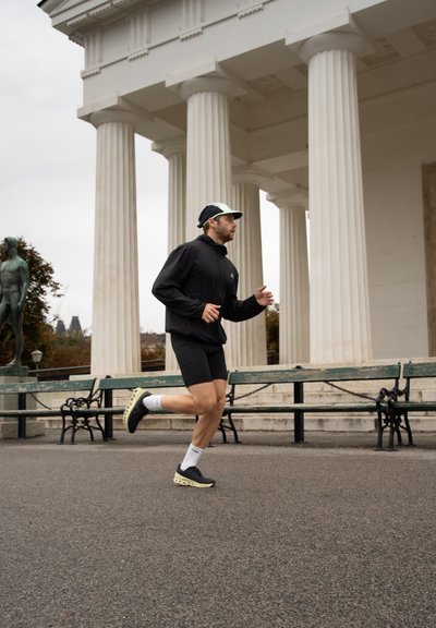Corredor con chaqueta negra con cremallera y pantalones cortos negros, lleva una gorra negra y verde, corre por un camino pavimentado al lado de bancos verdes y columnas.