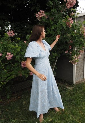 Vestido azul claro con bordado floral, escote en V, mangas cortas abullonadas, falda fluida, usado en un entorno al aire libre con plantas florecientes.