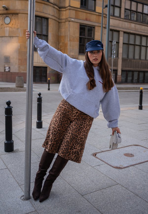 Femme portant un sweat à capuche gris, une jupe à imprimé léopard, des bottes noires et une casquette bleu marine, penchée sur le côté en tenant un poteau en métal dans un environnement urbain.