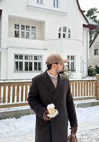 Homme en manteau sombre, gants marron et casquette tenant une tasse de café et un sac en cuir, debout sur un chemin enneigé devant une maison blanche avec une clôture en bois.