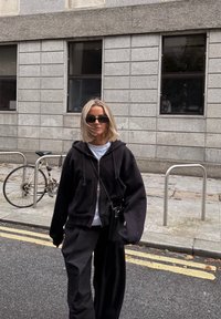 Felpa nera con zip sovrapposta a una maglietta bianca, abbinata a pantaloni neri larghi. Gli accessori includono occhiali da sole neri piccoli e una borsa.