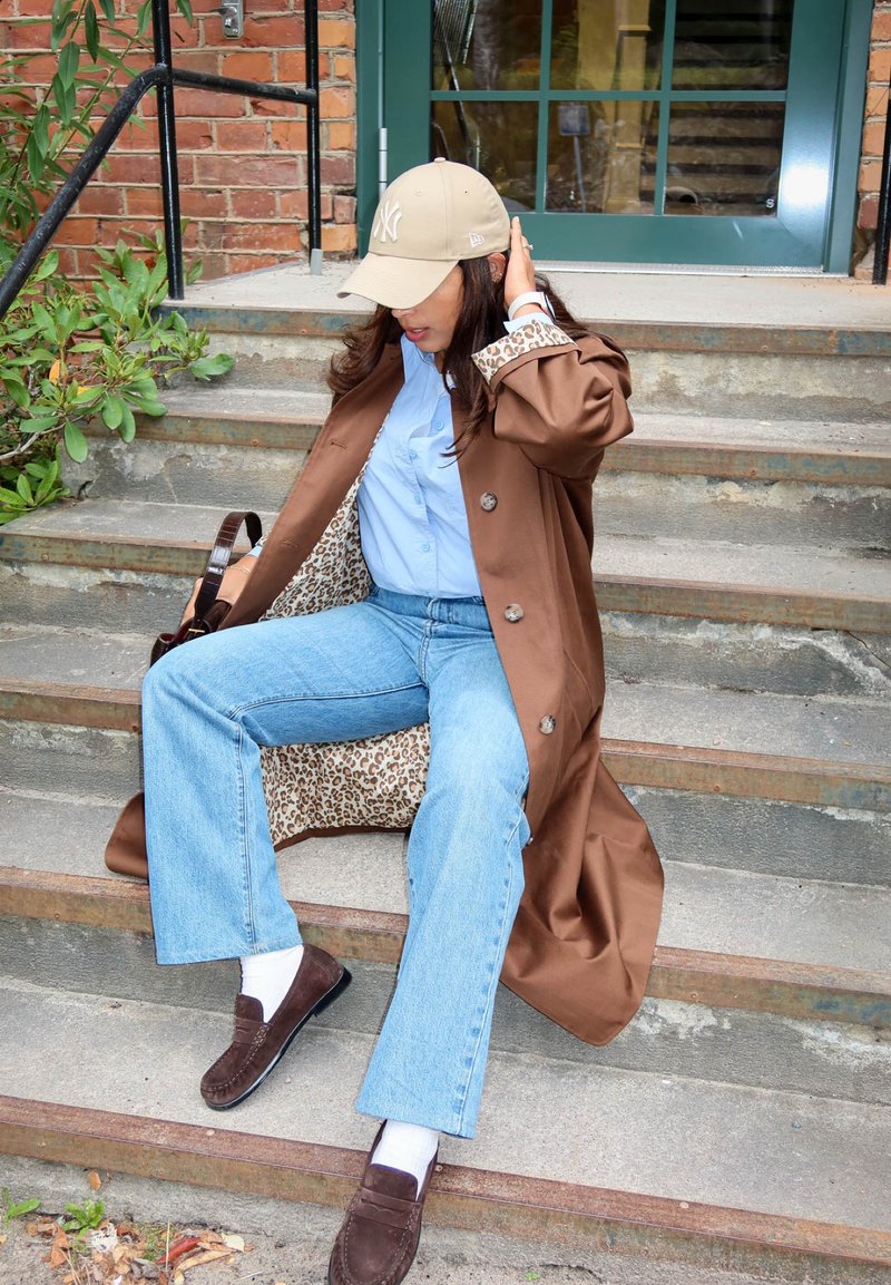 Mujer con abrigo marrón, camisa azul, vaqueros claros y gorra beige sentada en unas escaleras de hormigón al aire libre cerca de una puerta verde, arreglándose el pelo.