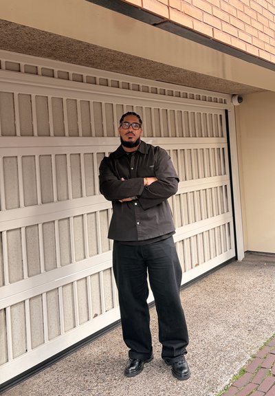 Hombre con gafas y barba, de brazos cruzados, vestido con ropa negra delante de una puerta de garaje cerrada y una pared de ladrillos.