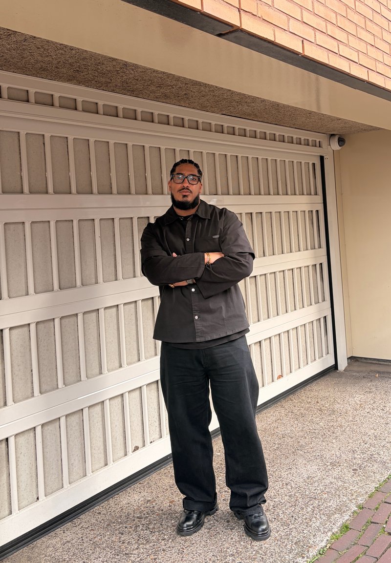 Homme avec lunettes et barbe, debout, bras croisés, portant des vêtements noirs devant une porte de garage fermée et un mur en briques.