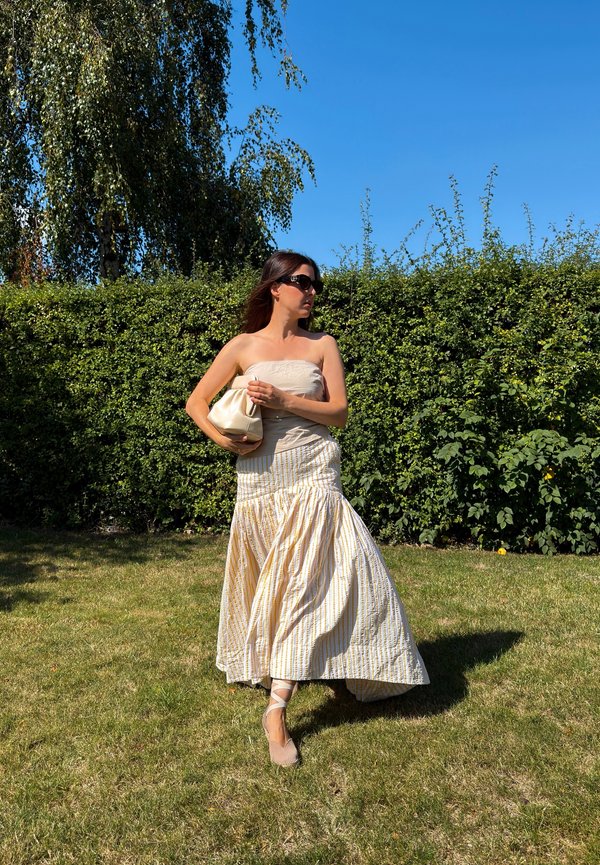 Femme en top sans bretelles et longue jupe marche sur l'herbe en tenant un petit sac clair, avec une haie verte et un ciel bleu en arrière-plan.