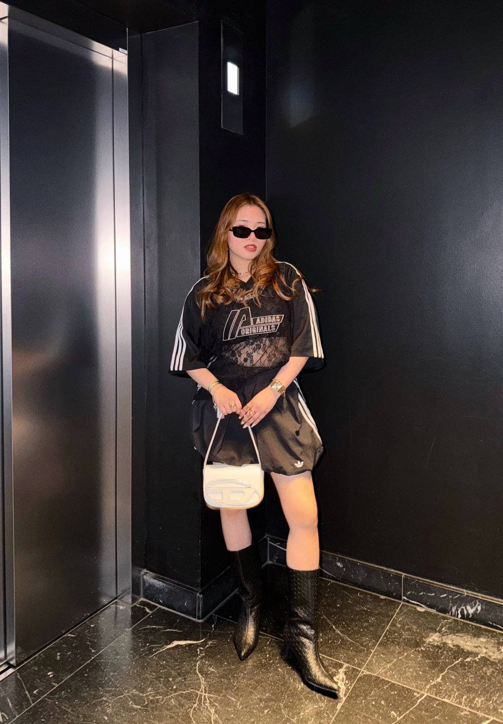 Chemise noire surdimensionnée Adidas avec accents en dentelle, associée à une jupe noire. Bottes noires montant jusqu'aux genoux et petit sac à main blanc. Fond sombre.