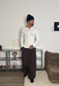 Chemise henley blanche à manches longues avec trois boutons, associée à un pantalon ample marron foncé. Bonnet noir, chaussures brillantes à faible contraste. Fond neutre.