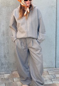 Femme portant un sweat à capuche gris surdimensionné et un pantalon large assorti, un foulard orange à motifs et des lunettes de soleil noires, debout contre un mur en béton.
