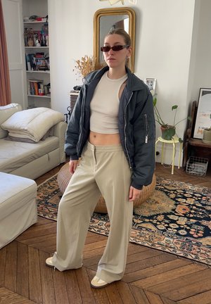Schwarze Pufferjacke, cropped weißes Top, beige weite Hosen und hellfarbige Schuhe. Accessoires umfassen schmale Sonnenbrillen und Ohrringe.