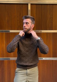 Homme ajustant le col d'un pull marron en maille torsadée, debout contre un mur en bois avec des barres métalliques horizontales.