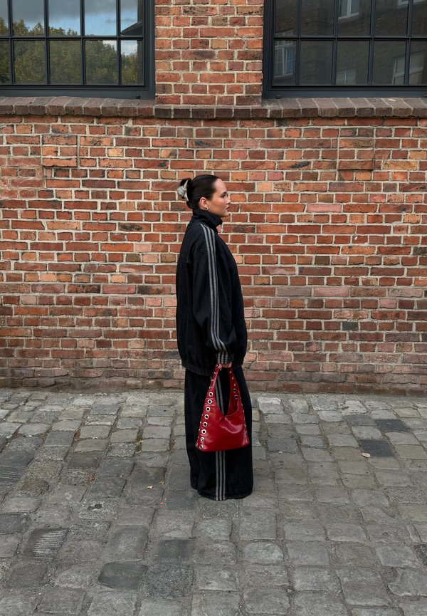 Femme debout de profil dans une rue pavée, portant un survêtement noir et tenant un sac à main rouge contre un mur de briques avec des fenêtres au-dessus.