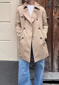Cappotto trench beige doppiopetto con spalline, grandi bottoni e tasche laterali, indossato sopra una maglietta bianca e jeans azzurri chiari.