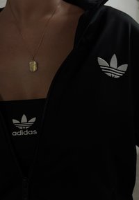 Artimiausias asmens vaizdas, vilkinčio juodą Adidas užtrauktuku striukę ir marškinėlius su baltais Adidas logotipais, ir auksinį stačiakampį pakabuką ant kaklo.
