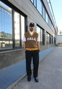 Knitted yellow and dark green striped vest featuring "YALE" text, worn over a white t-shirt. Paired with black tailored trousers and shiny black shoes.