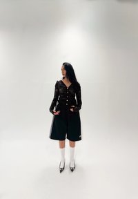 Zwart mesh top met lange mouwen en knopen, gecombineerd met zwarte knielange shorts met witte strepen, witte sokken en spitse hakken.