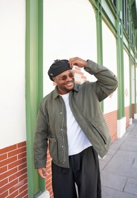 Homme en veste verte, chemise blanche, pantalon noir et béret ajuste son chapeau, souriant sur un trottoir urbain près d'un mur à cadre vert.