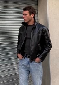 Homme debout, les mains dans les poches, portant une veste en cuir noire, une chemise noire et un jean bleu clair, devant un fond de volet métallique.