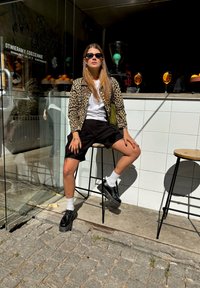 Casaco com padrão de leopardo sobre uma t-shirt branca, calções pretos e sapatos de plataforma pretos. Sentada num banco de bar em frente a um café com reflexos de vidro.
