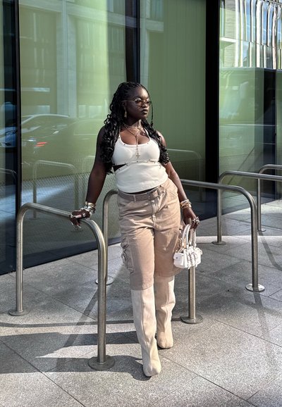 Modelo con una camiseta sin mangas blanca y pantalones cargo beige, apoyada en una barandilla metálica, vistiendo botas beige hasta la rodilla y sosteniendo un pequeño bolso blanco.