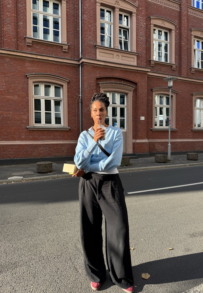 Hellblauer Pullover, schwarze weite Hose, hält eine Tasse mit einem Strohhalm, steht draußen vor einem Ziegelgebäude mit großen Fenstern.