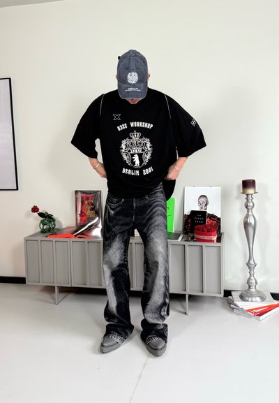 Camiseta negra de gran tamaño con gráfico y texto en blanco, combinada con jeans de patrón en negro y gris, y calzado gris. El escenario presenta muebles y decoración.