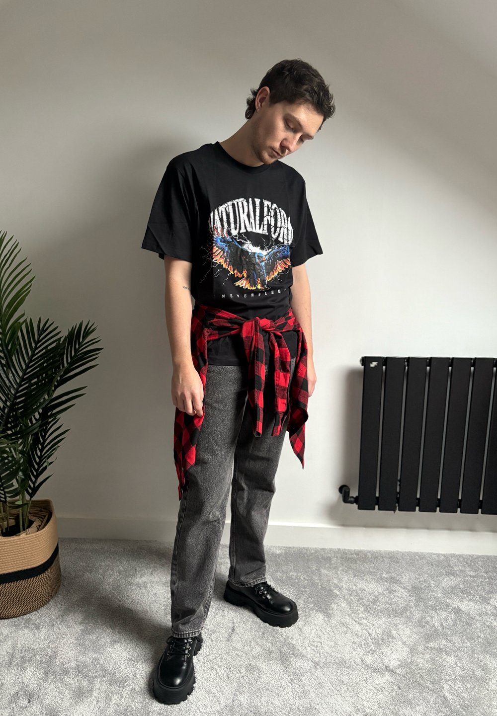 Jeune homme debout à l'intérieur portant un t-shirt graphique noir, une chemise à carreaux rouge nouée autour de la taille, un jean gris et des chaussures noires épaisses.