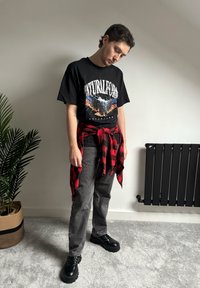Jeune homme debout à l'intérieur portant un t-shirt graphique noir, une chemise à carreaux rouge nouée autour de la taille, un jean gris et des chaussures noires épaisses.
