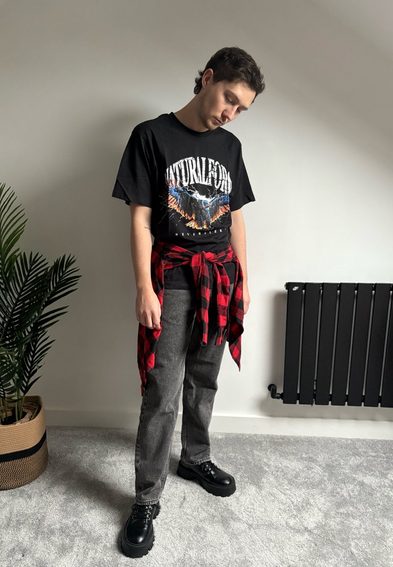 Jeune homme debout à l'intérieur portant un t-shirt noir avec un motif graphique, une chemise à carreaux rouge nouée autour de la taille, un jean gris et des chaussures noires épaisses.