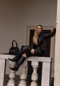 Tamna smeđa kožna jakna i suknja, crne cipele na visoku petu, sjedi na kamenoj ivici, drži crnu torbu s zlatnim detaljima.